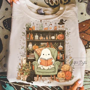 May include: A light beige sweatshirt featuring a whimsical Halloween-themed illustration. A friendly ghost reads a book in a green armchair in front of a bookshelf filled with potion bottles, pumpkins, and books. A black cat and pumpkins are at the bottom.