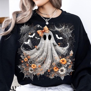 Puede incluir: Sudadera negra con un dise&ntilde;o de fantasma de Halloween. El fantasma tiene un lazo, rodeado de telara&ntilde;as, murci&eacute;lagos y flores naranjas, blancas y grises. Un gr&aacute;fico festivo para la temporada.