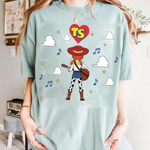 Puede incluir: Una camiseta de manga corta en color verde salvia claro con el gr&aacute;fico de una vaquera de pelo rojo y sombrero tocando la guitarra. El dise&ntilde;o incluye un coraz&oacute;n rojo con las letras TS, nubes blancas, estrellas amarillas y notas musicales azules.