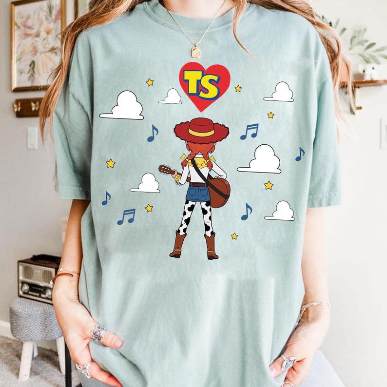 Puede incluir: Una camiseta de manga corta en color verde salvia claro con el gr&aacute;fico de una vaquera de pelo rojo y sombrero tocando la guitarra. El dise&ntilde;o incluye un coraz&oacute;n rojo con las letras TS, nubes blancas, estrellas amarillas y notas musicales azules.