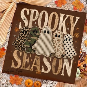Può includere: Un cartello rettangolare marrone con la scritta SPOOKY SEASON in lettere bianche dall'aspetto usurato. Cinque sagome di tessuto a forma di fantasma sono disposte al centro, con motivi leopardati, mimetici, a righe verticali, floreali e a pois.