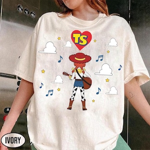 Puede incluir: Una camiseta color marfil con el gr&aacute;fico de una vaquera de dibujos animados con pelo rojo y sombrero, tocando la guitarra. Sobre el personaje hay un coraz&oacute;n rojo con las letras amarillas TS. El dise&ntilde;o incluye nubes blancas, estrellas y notas.