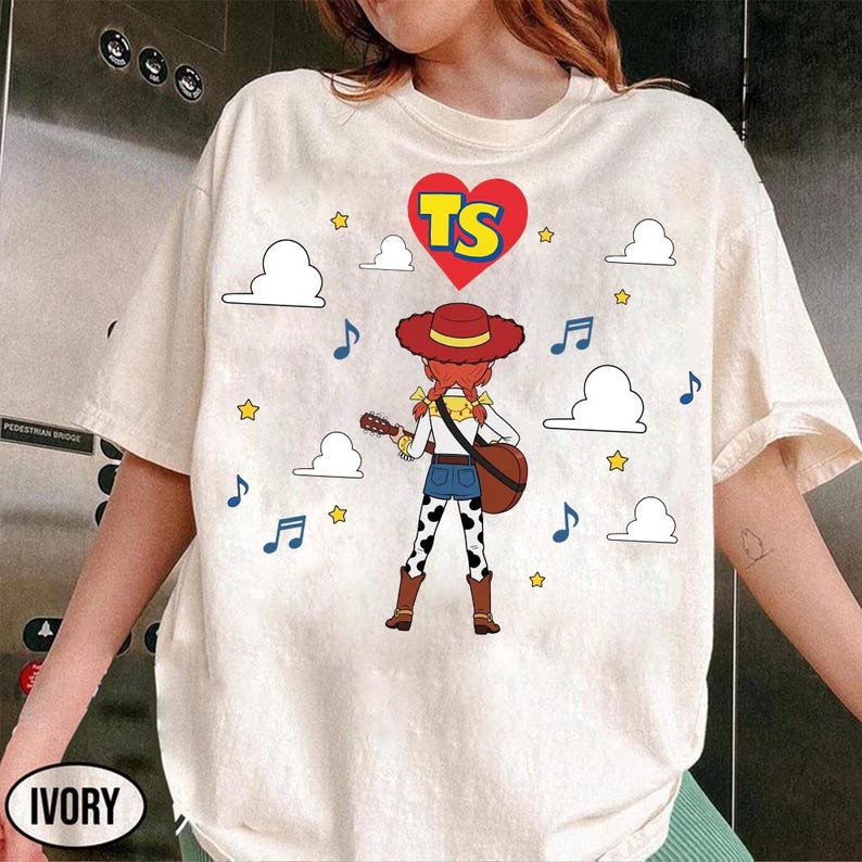 Puede incluir: Una camiseta color marfil con el gr&aacute;fico de una vaquera de dibujos animados con pelo rojo y sombrero, tocando la guitarra. Sobre el personaje hay un coraz&oacute;n rojo con las letras amarillas TS. El dise&ntilde;o incluye nubes blancas, estrellas y notas.