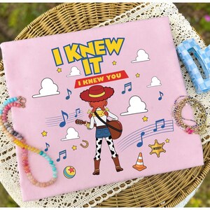 May include: A light pink fabric square featuring a graphic of a cowgirl character with red hair and a hat playing a guitar. The text reads I KNEW IT and I KNEW YOU. The design includes blue musical notes, white clouds, yellow stars, a sheriff badge, and a ball.