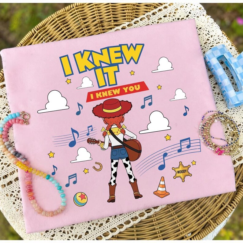 May include: A light pink fabric square featuring a graphic of a cowgirl character with red hair and a hat playing a guitar. The text reads I KNEW IT and I KNEW YOU. The design includes blue musical notes, white clouds, yellow stars, a sheriff badge, and a ball.