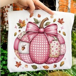 May include: A white t-shirt featuring a large, central graphic of a patchwork-style pumpkin in shades of pink. The pumpkin is decorated with a prominent pink bow and surrounded by small illustrations of autumn leaves, acorns, and corn cobs.