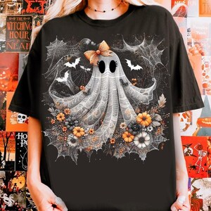 Puede incluir: Camiseta negra con un dise&ntilde;o caprichoso de fantasma con lazo, rodeado de telara&ntilde;as, murci&eacute;lagos y flores en naranja, amarillo y gris. El dise&ntilde;o tiene un tema de Halloween.