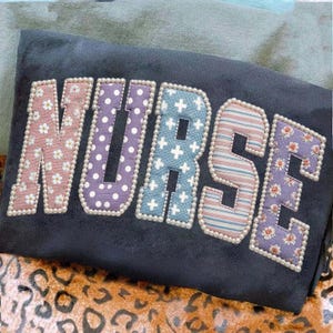 Könnte beinhalten: Ein dunkelgraues Sweatshirt mit dem Wort "NURSE" auf der Vorderseite. Die Buchstaben sind in verschiedenen Mustern gehalten, darunter Blumen, Punkte und Streifen, und sind mit kleinen weißen Perlen umrandet.