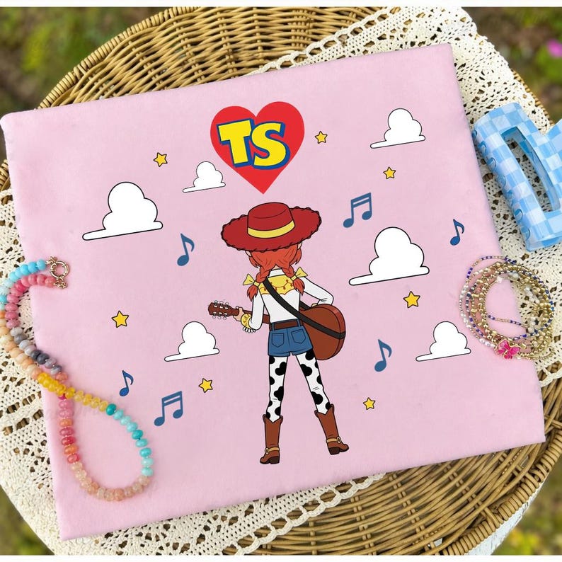 Puede incluir: Una superficie de tela rosa claro con una ilustraci&oacute;n de dibujos animados de una vaquera con cabello rojo, un sombrero rojo y una guitarra. El dise&ntilde;o incluye un coraz&oacute;n rojo con las letras TS, nubes blancas, estrellas amarillas y notas musicales azules.
