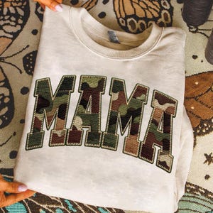 Op de afbeelding: Een crèmekleurige sweatshirt met het woord "MAMA" in grote blokletters. De letters zijn gevuld met een camouflagepatroon in groen, bruin en beige, en omrand met donkergroene stiksels.