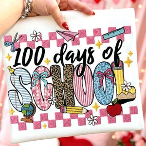 May include: White square fabric with the text "100 days of SCHOOL" in a variety of colorful patterns and designs. The design includes a globe, scissors, a pencil, a ruler, a paper airplane, and an apple. Pink and white checkered border.