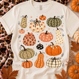 May include: A cream-colored short-sleeve t-shirt featuring a graphic print of various pumpkins in different colors, patterns, and textures, including polka dots, leopard print, stripes, and floral motifs, accented with small stars and dots.