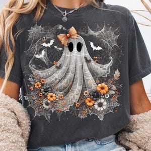 May include: Charcoal grey t-shirt with a Halloween ghost graphic. The ghost has a bow and is set against spiderwebs, bats, and orange, black, and white floral elements. A spooky graphic tee.