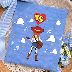 Puede incluir: Un cuadrado de tela azul claro con un personaje de dibujos animados de pelo rojo, sombrero rojo y pantalones con manchas de vaca tocando la guitarra. El dise&ntilde;o incluye nubes blancas, estrellas amarillas, notas musicales y un coraz&oacute;n rojo con TS.