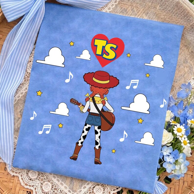 Puede incluir: Un cuadrado de tela azul claro con un personaje de dibujos animados de pelo rojo, sombrero rojo y pantalones con manchas de vaca tocando la guitarra. El dise&ntilde;o incluye nubes blancas, estrellas amarillas, notas musicales y un coraz&oacute;n rojo con TS.