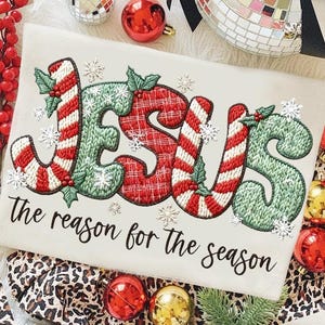 May include: Cream-colored Christmas-themed sweatshirt with the word "JESUS" embroidered in red, green, and white candy cane and plaid patterns. The text "The reason for the season" is below the word "JESUS". Decorative snowflakes and holly accents are also present.