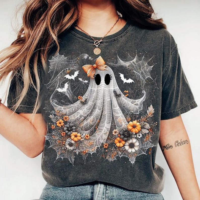 Puede incluir: Camiseta gris oscuro con un dise&ntilde;o de fantasma. El fantasma tiene un lazo, rodeado de telara&ntilde;as, murci&eacute;lagos y flores naranjas, grises y blancas. Una camiseta gr&aacute;fica con tem&aacute;tica de Halloween.