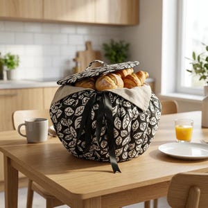 May include: A round bread basket with a black and white leaf design, filled with bread. The basket has a lid and is tied with black ribbons. A cup of coffee, a glass of orange juice, and a plate are on the wooden table.