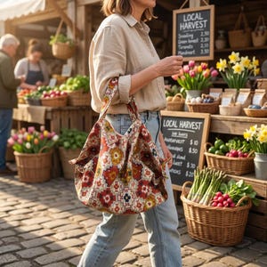 Puede incluir: Una mujer lleva un bolso tote grande con un diseño geométrico rojo, amarillo y azul. El bolso tiene asas a juego. El fondo muestra un mercado de agricultores con cestas de productos y flores, y un letrero que dice "LOCAL HARVEST".