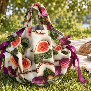 May include: A fabric bag featuring a repeating pattern of sliced and whole purple figs with green leaves on a cream background. The bag has two top handles and is cinched at the sides with dark purple ribbon ties, creating a gathered, rounded shape.