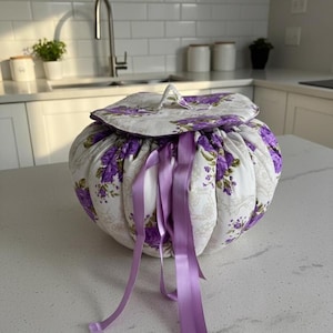 Könnte beinhalten: Ein runder, gesteppter Stoffbehälter mit Deckel, mit einem Blumenmuster in Lila und Weiß. Der Behälter ist oben gerafft und mit langen, lavendelfarbenen Bändern gebunden. Der Hintergrund ist eine Küche.