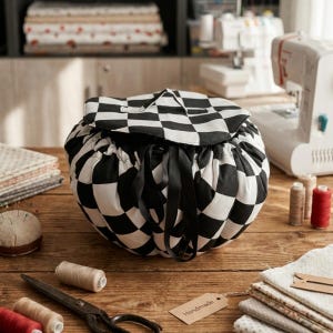 May include: A black and white chequered drawstring bag with black ribbon ties. The round bag is on a wooden surface with sewing supplies, including thread spools, scissors, and fabric scraps. A tag that says "Handmade" is attached.