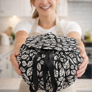 Könnte beinhalten: Eine runde Reisetasche mit einem schwarz-weißen Blättermuster. Die Tasche wird mit schwarzen Bändern offen gehalten. Die Tasche wird von einer Person gehalten, die ein weißes Oberteil und eine Schürze trägt.