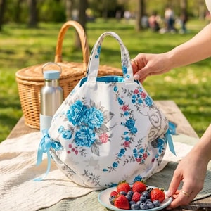 Może przedstawiać: Biała kwiatowa torba piknikowa z niebieskimi i różowymi kwiatami, niebieskimi wstążkami i pasującym niebieskim wnętrzem. Wiklinowy kosz piknikowy i talerz truskawek i jagód znajdują się na drewnianym stole piknikowym.