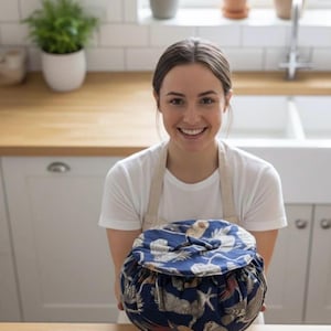 Pode incluir: Saco redondo azul marinho com cordão com um padrão de pássaros e conchas. O saco é segurado em frente a uma pessoa em uma cozinha. O saco tem cerca de 25 cm de diâmetro.