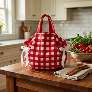 Puede incluir: Una bolsa de picnic a cuadros rojos y blancos con asas rojas y lazos de cinta blancos. La bolsa está sobre una encimera de madera en un entorno de cocina. También se ve un cuenco de verduras frescas y cucharas de madera.