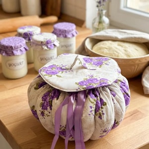 Puede incluir: Un recipiente redondo de tela con un estampado floral en morado y blanco, con cierre de cordón y cintas moradas. El recipiente tiene tapa. Frascos de yogur con tapas moradas están en el fondo, sugiriendo un tema de cocina.