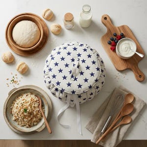 Puede incluir: Un calentador de pan blanco con un patrón de estrellas azul marino, rodeado de cuencos de madera con masa y panecillos, una tabla de cortar con bayas y yogur, y un cuenco de arroz. También hay utensilios de cocina e ingredientes.