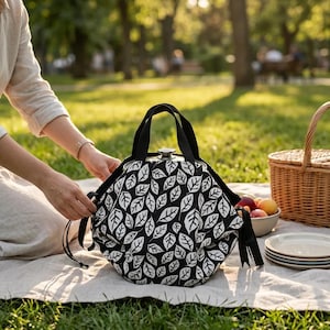 Puede incluir: Una cesta de picnic con estampado de hojas en blanco y negro con asas negras. La cesta está sobre una manta blanca con una cesta de picnic, platos y un cuenco de fruta en el fondo. La escena está ambientada al aire libre en un parque.