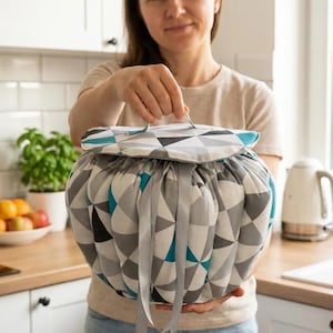 May include: A round fabric storage bag with a handle, featuring a geometric pattern in grey, teal, and white. The bag is held by a person in a kitchen. It is tied with grey ribbons.