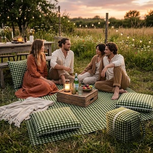 Op de afbeelding: Een groen-wit geblokt picknickkleed met bijpassende vierkante vloerkussens en een kubusvormige koeltas. Een houten krat dient als tafel met daarop een glazen fles, twee drinkglazen en een schaal met fruit op het kleed.