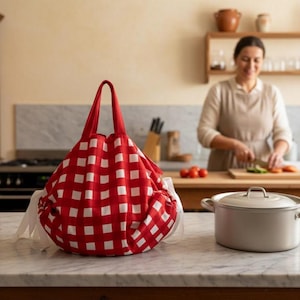 Può includere: Una borsa a coulisse a scacchi rossi e bianchi con un manico abbinato è appoggiata su un piano di lavoro in marmo. Una pentola d'argento con coperchio è accanto alla borsa. Sullo sfondo, una persona sta preparando il cibo in una cucina.