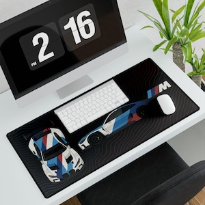 May include: Black rectangular desk mat featuring two stylized race cars in blue, red, and white. The mat has a black background with a wavy pattern and the BMW logo. A white keyboard and mouse are on the mat.