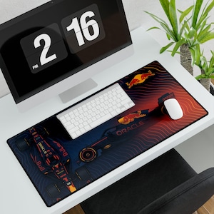 May include: A desk setup featuring a large mouse pad with a Formula 1 racing car design in navy blue, red, and yellow. The mouse pad has the Oracle and Red Bull logos. A white keyboard and mouse sit on the pad, with a computer monitor displaying the time.