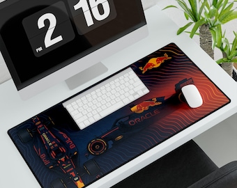 May include: A desk setup featuring a large mouse pad with a Formula 1 racing car design in navy blue, red, and yellow. The mouse pad has the Oracle and Red Bull logos. A white keyboard and mouse sit on the pad, with a computer monitor displaying the time.