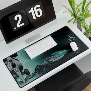 May include: A black desk mat with a Formula 1 race car design in teal and white. The mat features the Mercedes-Benz logo and the words "AMG PETRONAS". A white keyboard and mouse are on the mat, with a computer monitor in the background.