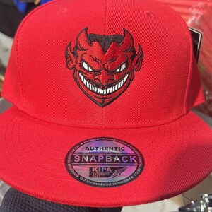 May include: A red snapback hat featuring a black devil face design. The hat has a flat brim and an adjustable snap closure. The hat has the words "AUTHENTIC SNAPBACK" printed on the front.