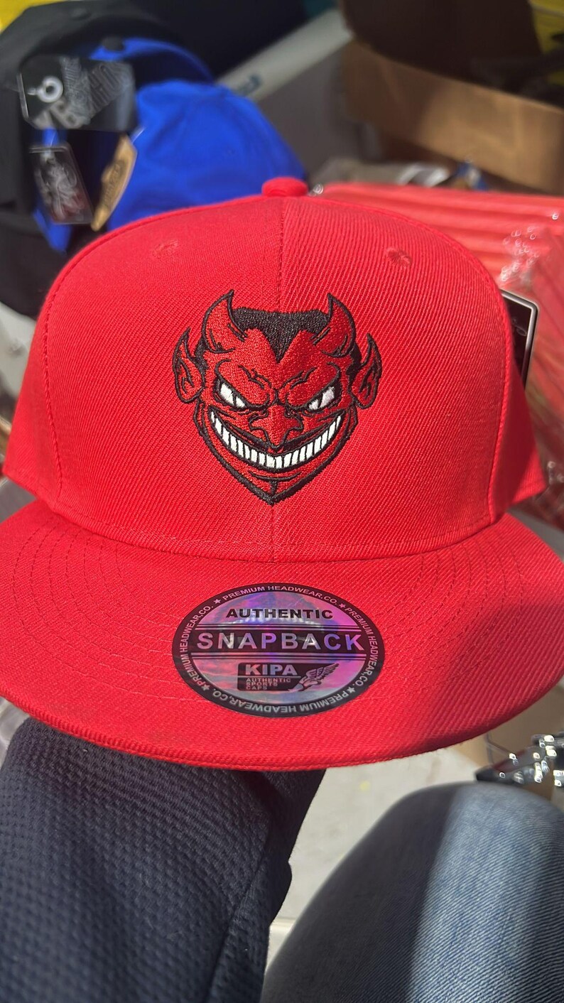May include: A red snapback hat featuring a black devil face design. The hat has a flat brim and an adjustable snap closure. The hat has the words "AUTHENTIC SNAPBACK" printed on the front.