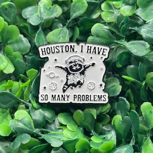 Può includere: Spilla smaltata bianca e nera con la scritta "HOUSTON, I HAVE" in alto e "SO MANY PROBLEMS" in basso. La spilla raffigura un procione con un casco spaziale a braccia aperte, circondato da stelle e pianeti.