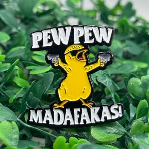 Puede incluir: Un pin de esmalte de pollo de dibujos animados amarillo con las palabras "PEW PEW" arriba y "MADAF*KAS!" abajo. El pollo sostiene dos pistolas grises. El pin es blanco y negro.
