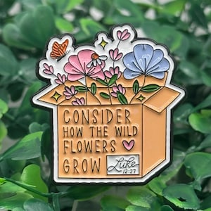 May include: A colorful enamel pin featuring a box overflowing with pink and blue flowers, a butterfly, and the text "Consider How The Wild Flowers Grow" with a Bible verse reference. The pin has a black outline.