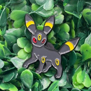 Może przedstawiać: Czarny emaliowany pin przedstawiający stylizowaną istotę z dużymi uszami i ogonem. Pin ma żółte akcenty na uszach i ogonie, z żółtymi i czerwonymi detalami na oczach i ciele. Pin jest umieszczony na tle zielonych liści.