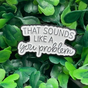 May include: A white enamel pin with the phrase "THAT SOUNDS LIKE A you problem" in black lettering. The pin has a black border and is set against a background of green foliage.