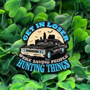 Puede incluir: Pin esmaltado con un coche negro con detalles azules, una silueta de ciudad y el texto "GET IN LOSER WERE SAVING PEOPLE HUNTING THINGS" en letras azules. El pin está sobre un fondo de follaje verde.