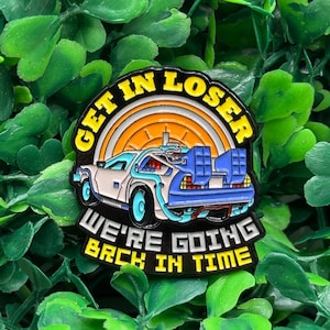 May include: A colorful enamel pin featuring a stylized DeLorean time machine with the text "GET IN LOSER" and "WE'RE GOING BACK IN TIME". The car is white and blue, set against a yellow and orange background, with black text and border.