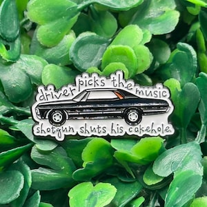 Puede incluir: Un pin de esmalte negro con forma de coche con el texto "driver picks the music" encima del coche y "shotgun shuts his cakehole" debajo. El pin tiene un borde plateado y está sobre un fondo de hojas verdes.